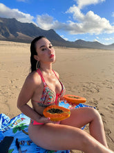 Laden Sie das Bild in den Galerie-Viewer, Model in a colorful bikini sitting on the beach holding bowls of papaya with mountains in the background.