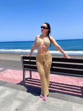 Load image into Gallery viewer, Model wearing luxe crochet trousers in gold by the beach, showcasing summer fashion and a flattering fit.