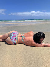 Load image into Gallery viewer, Model wearing a hand beaded ruched bikini lying on the beach, showcasing vibrant patterns and a relaxed pose.