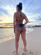 Load image into Gallery viewer, Model wearing a hand beaded Brazilian bikini on the beach at sunset, showcasing vibrant patterns and handcrafted details.