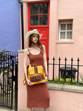 Load image into Gallery viewer, Fashionable woman in a brown dress holding a yellow handbag against a colorful building backdrop.