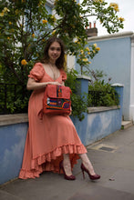 Load image into Gallery viewer, Woman in an orange dress sitting with a handcrafted tangerine leather rucksack with Mola applique.