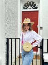 Load image into Gallery viewer, Woman wearing a handmade Iraca palm seahorse bag with a hat, standing by a red door in casual attire.