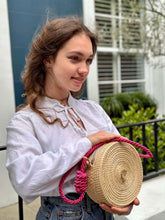 Load image into Gallery viewer, Young woman holding a handmade Iraca palm seahorse canteen bag with pink braided handle outside.