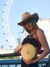 Load image into Gallery viewer, Model showcasing a handmade Iraca palm seahorse canteen bag with a stylish hat near a city landmark.