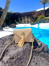 Load image into Gallery viewer, Handwoven Iraca palm leaf shoulder bag with tassel detail, placed on a rocky surface by a blue swimming pool.