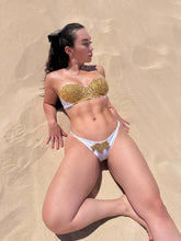Cargar imagen en el visor de la galería, Model wearing Nina hand-beaded white bikini with gold accents, lounging on sandy beach.