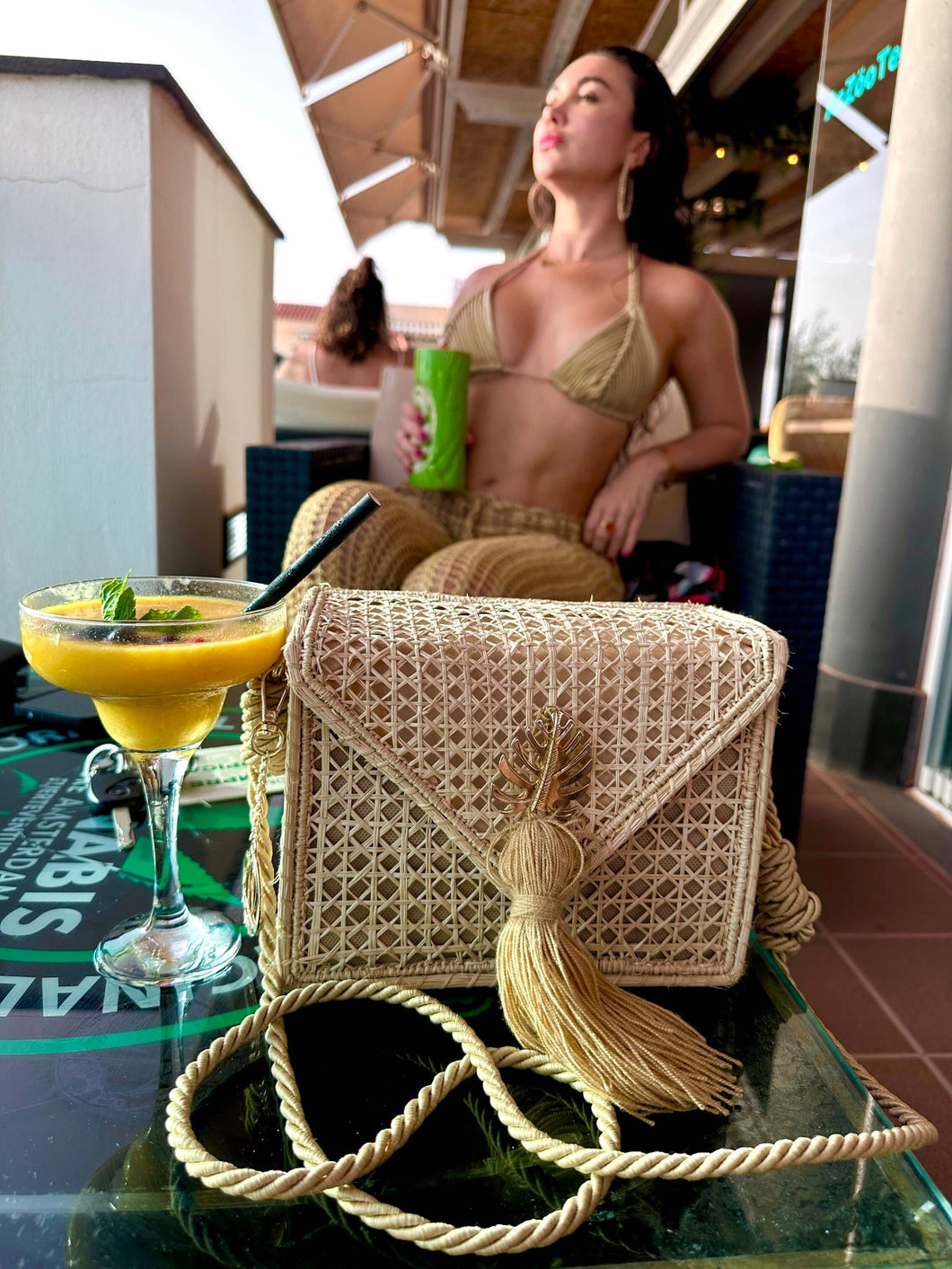 Handwoven Iraca palm leaf shoulder bag styled with a cocktail at a sunny outdoor café.